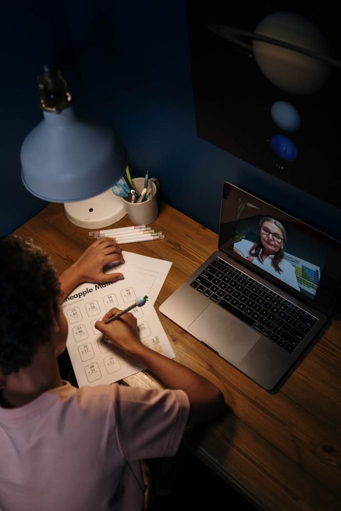 overhead-shot-of-a-student-doing-his-homework-while-listening-to-an-online-class-7014770 Child engaged in online learning session with a teacher via laptop.