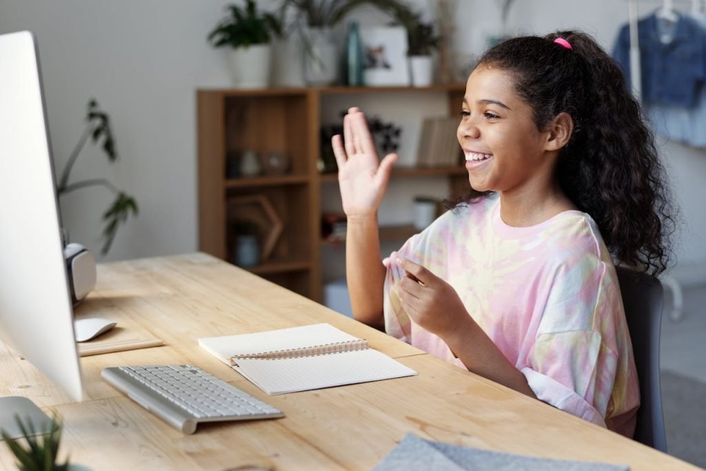 woman-in-pink-shirt-sitting-by-the-table-while-smiling-4143791 A happy girl waves during an online class at home, sitting at a desk with a computer.